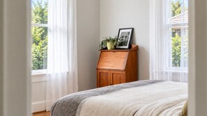 Una camera da letto con un letto, un comodino in legno con una pianta e un portafoto, e una finestra con tende leggere.