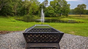 A fire pit with a metal grate, surrounded by gravel, in a park with a pond and trees. - Green Mountain Retreat: Pocono Suites (Pleasant Mount)