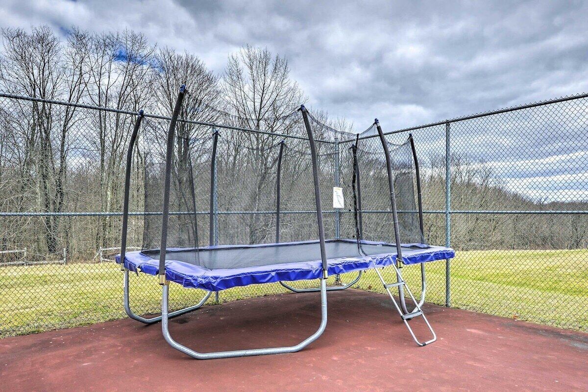 A trampoline enclosed by a chain-link fence with a red surface, surrounded by a grassy area and leafless trees.