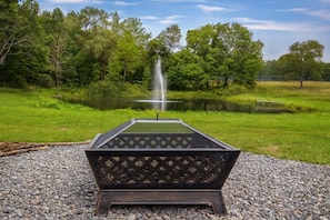 A fire pit with a metal grate, surrounded by gravel, in a park with a pond and trees.