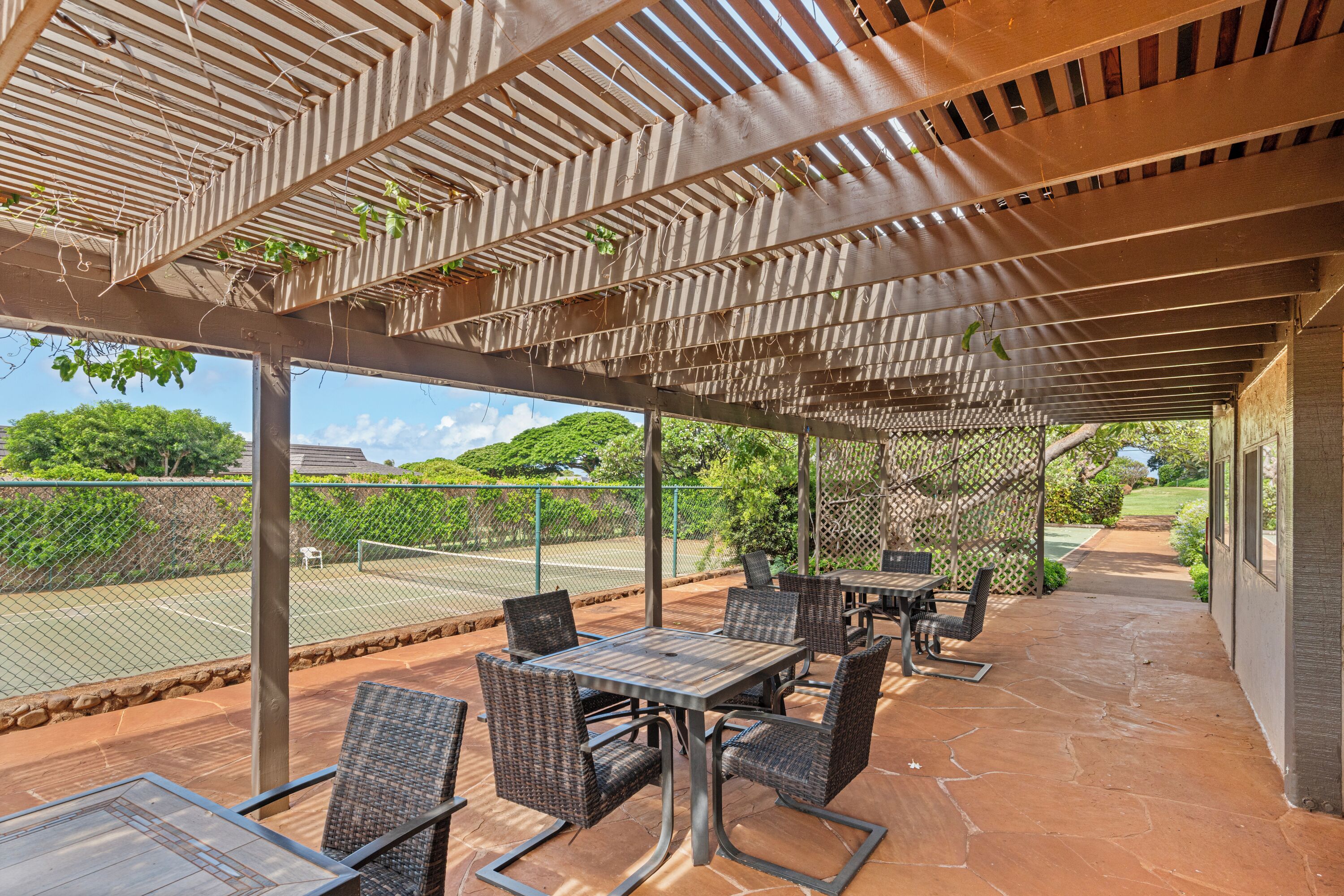 An outdoor patio with tables and chairs, a tennis court in the background, and a covered area with a pergola.