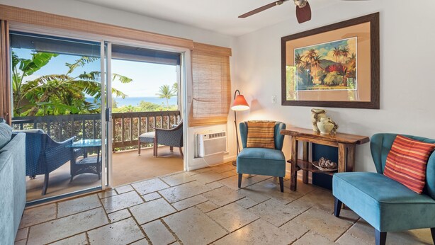 A room with a sliding glass door leading to a balcony with a view of the ocean. There is a ceiling fan, a table with chairs, and a sofa with a cushion. - Kaanapali Plantation By Chase N Rainbows (Lahaina)