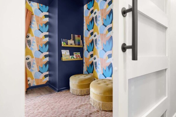 A room with a dark blue accent wall, a patterned curtain, and a shelf with books and decorative items. There are two round, beige ottomans in the foreground.