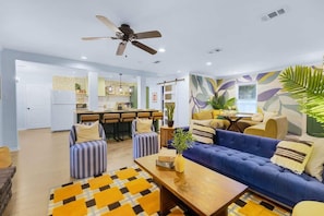 A modern living room with a blue sofa, striped armchairs, and a wooden coffee table. A ceiling fan is visible, and the room has a large window with a view of the kitchen.