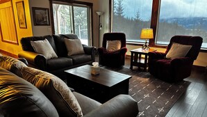 Ein gemütliches Wohnzimmer mit dunklem Holzboden, Ledersofas und Sesseln. Ein Couchtisch mit Taschentuchbox, eine Lampe auf einem Beistelltisch und ein großes Fenster mit Blick auf Bäume und Berge.