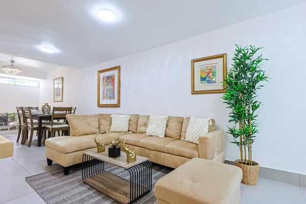 Ein Wohnzimmer mit einem beigefarbenen Sofa, einem Couchtisch und einem Essbereich mit Stühlen und einem Tisch. An der Wand hängen zwei gerahmte Kunstwerke und ein Blumentopf steht dort.