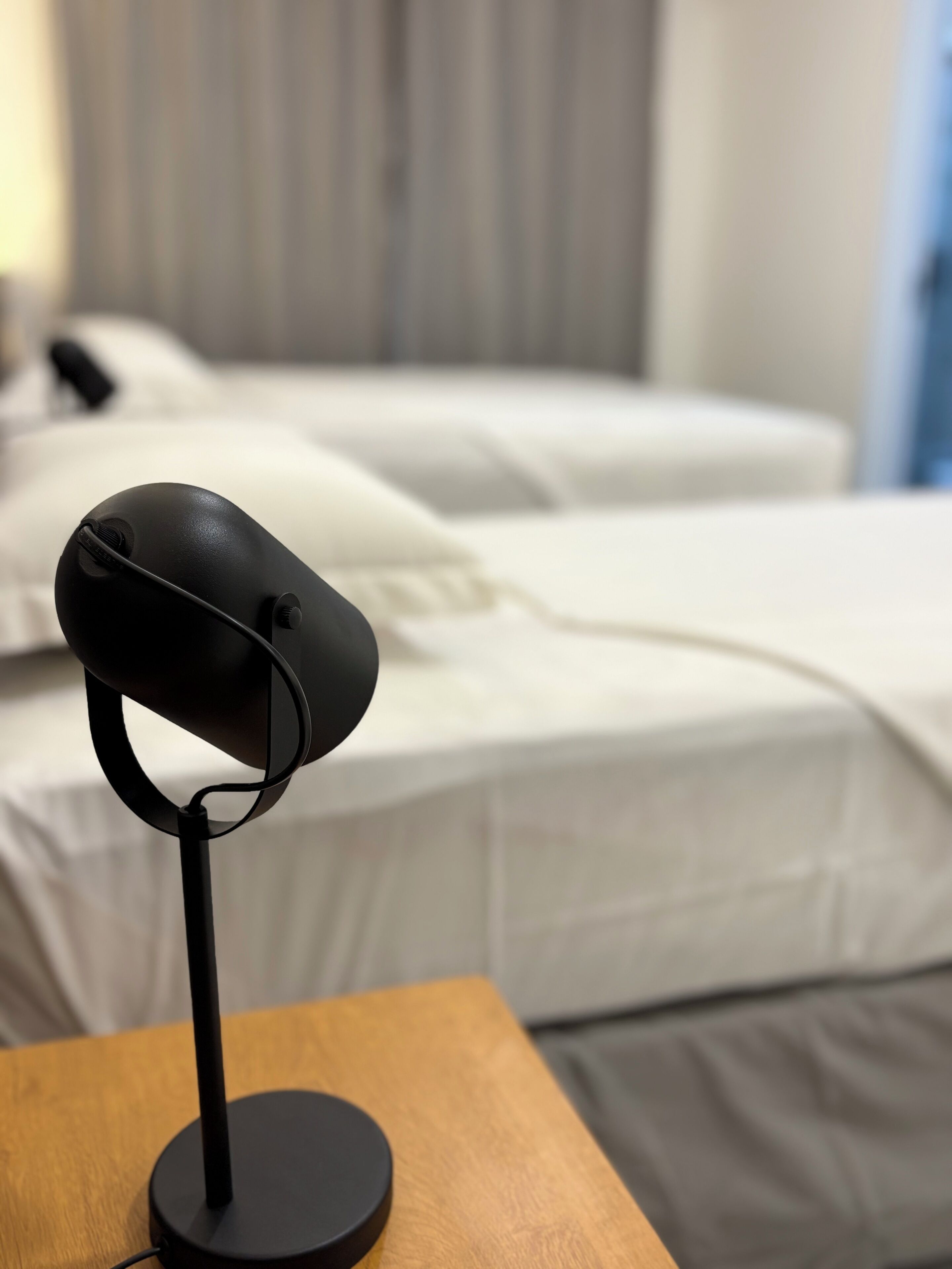 Een bed met wit beddengoed, een zwart object op een houten tafel en een gordijn op de achtergrond.