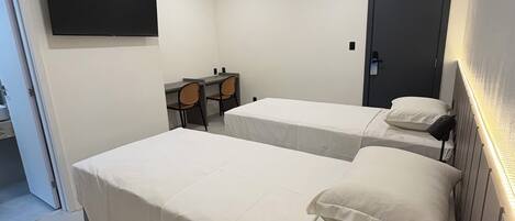 A modern hotel room with two single beds, a desk with a chair, a flat-screen TV mounted on the wall, and a door.