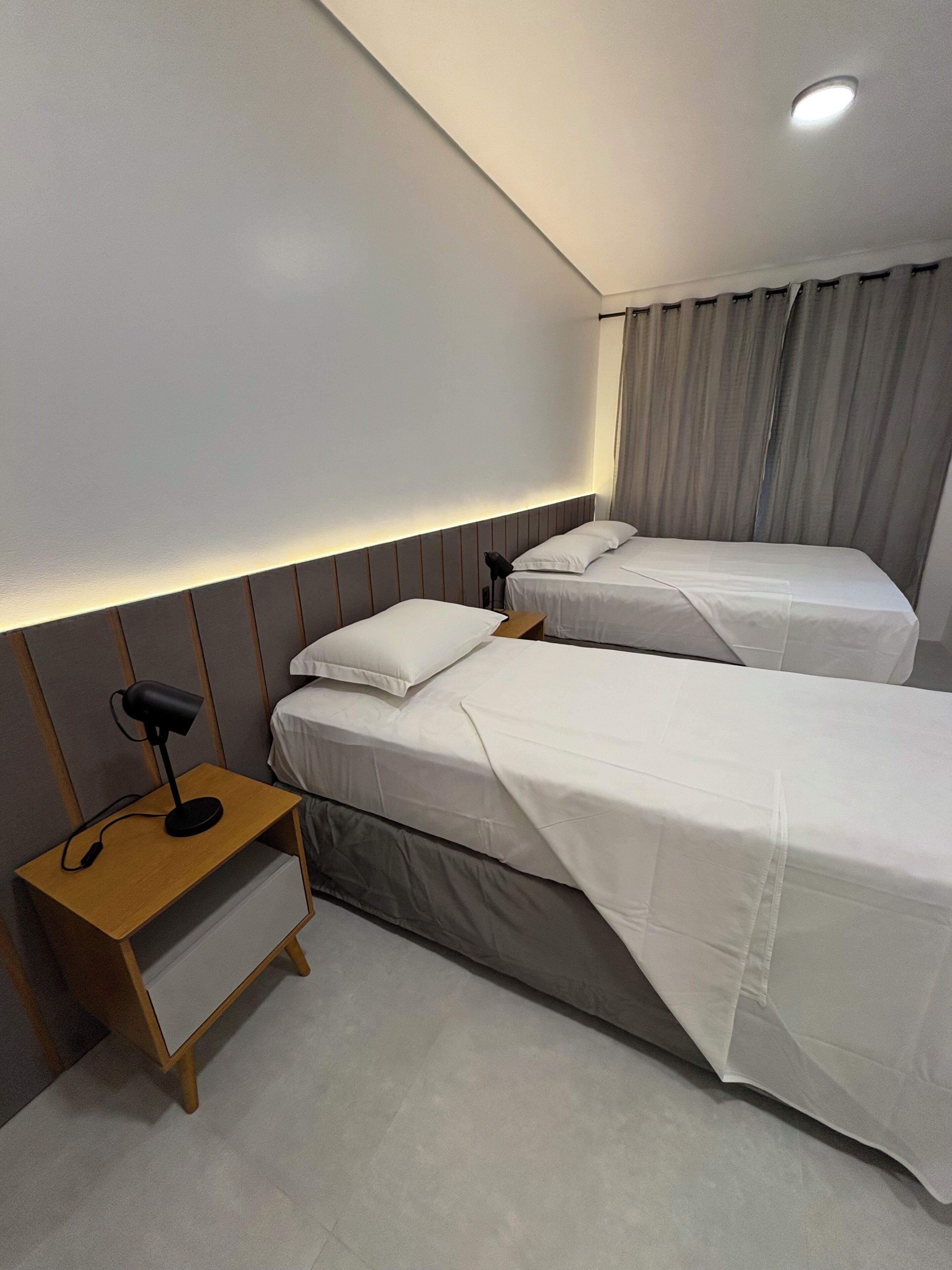 A modern hotel room with two single beds, a wooden nightstand with a lamp, and a ceiling light.