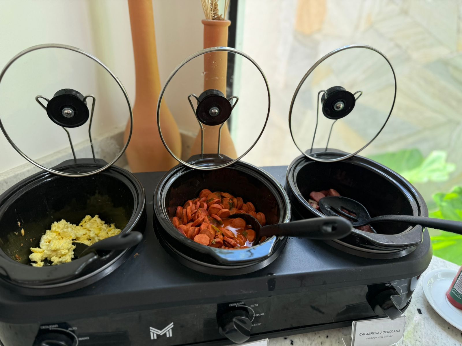 En ångkokare med tre behållare med lock, varav en innehåller rörägg, en innehåller korv och en innehåller sås.
