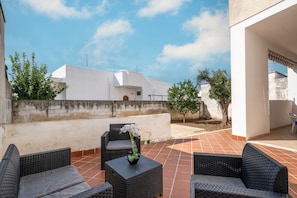 A patio with wicker furniture, a potted plant, and a white building in the background.