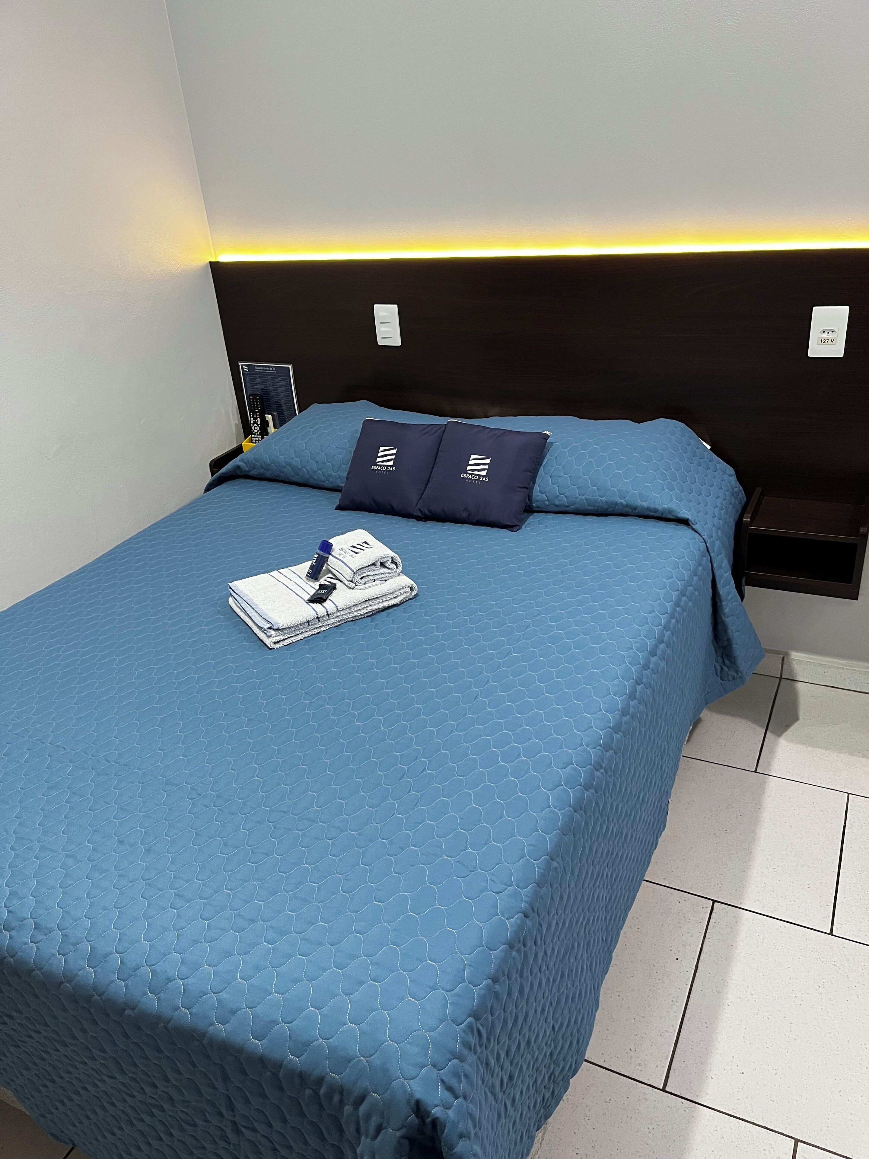 A neatly made bed with a blue quilt, two matching pillows, and a folded towel on the bed.