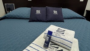 Habitación de hotel con cama, dos almohadas con el logo del hotel, un control remoto y una toalla con el logo del hotel.