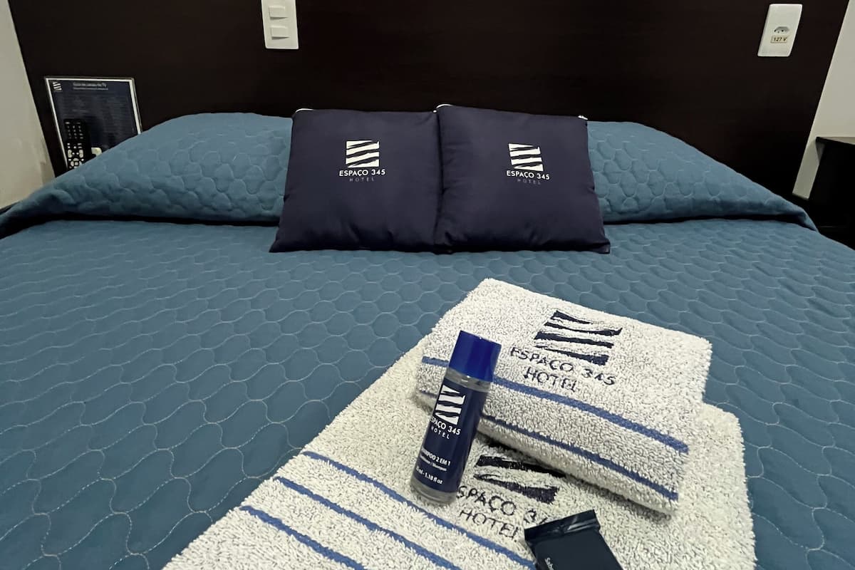 Une chambre d’hôtel comprenant un lit, deux oreillers ornés du logo de l’hôtel, une télécommande et une serviette également marquée du logo de l’hôtel.