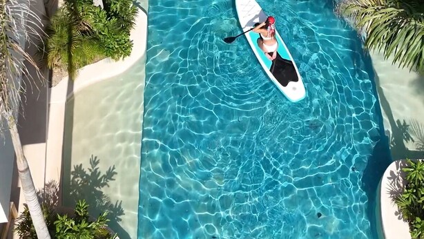 A clear blue pool with a paddleboarder, surrounded by palm trees and a multi-level building. - COCAY LOFTS BY SANDY POWERFUL (Tulum)