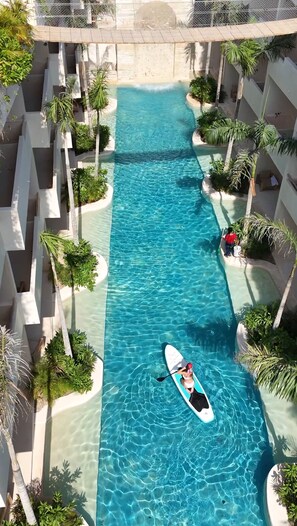 A clear blue pool with a paddleboarder, surrounded by palm trees and a multi-level building. - COCAY LOFTS BY SANDY POWERFUL (Tulum)