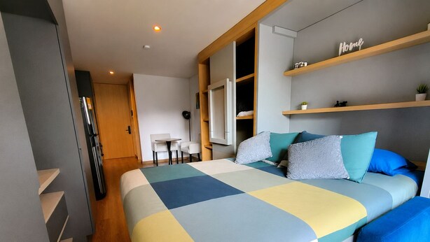 Une chambre moderne avec un grand lit, un bureau et une chaise. Il y a des étagères au mur et une porte qui donne sur une autre pièce.