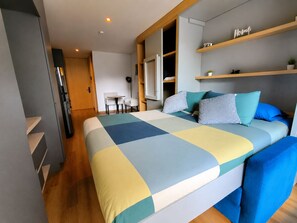 Une chambre moderne avec un grand lit, un bureau et une chaise. Il y a des étagères au mur et une porte qui donne sur une autre pièce.
