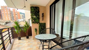Un balcon avec une table ronde en métal et deux chaises en fil de fer noir, des plantes en pot et une vue sur les immeubles à travers de grandes fenêtres.