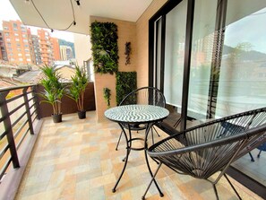 Een balkon met een ronde metalen tafel en twee zwarte draadstoelen, bloempotten en een uitzicht op gebouwen door grote ramen.