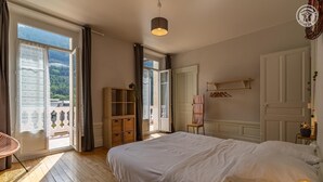 Um quarto com uma cama grande, um guarda-roupa de madeira, uma cadeira, uma planta pendente e vista para uma varanda com grade.