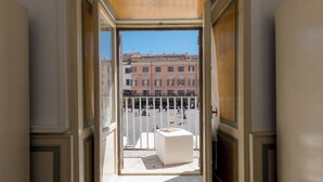 Une vue depuis un balcon, avec un siège blanc doté d’un coussin et une table, donnant sur une place bordée de bâtiments et sous un ciel dégagé.