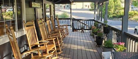 A wooden deck with rocking chairs and a table, overlooking a street with parked cars and greenery.