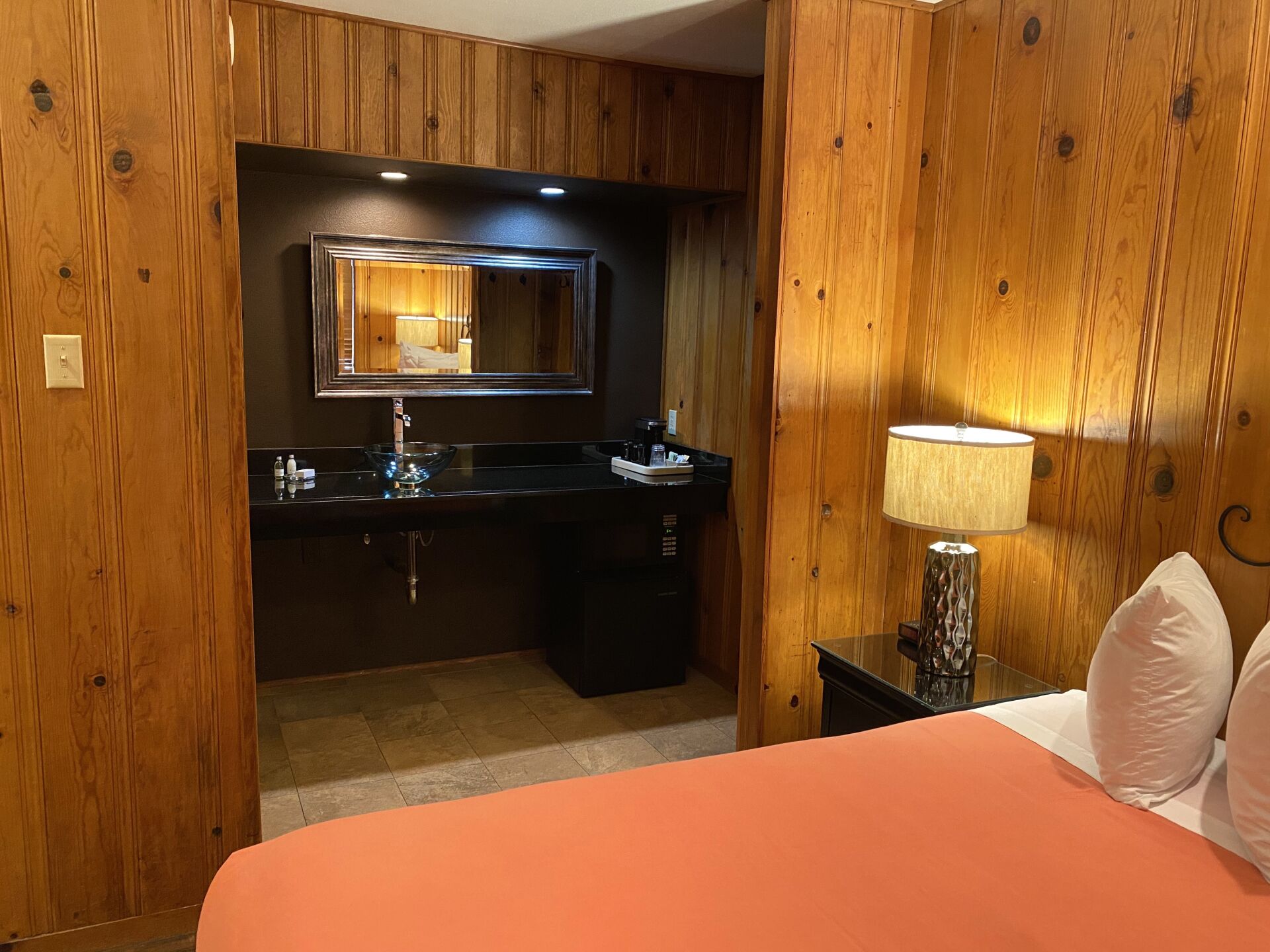 Habitació d'hotel amb paret de fusta, un tocador negre amb mirall, un rentapans i una taula de nit amb una làmpada.