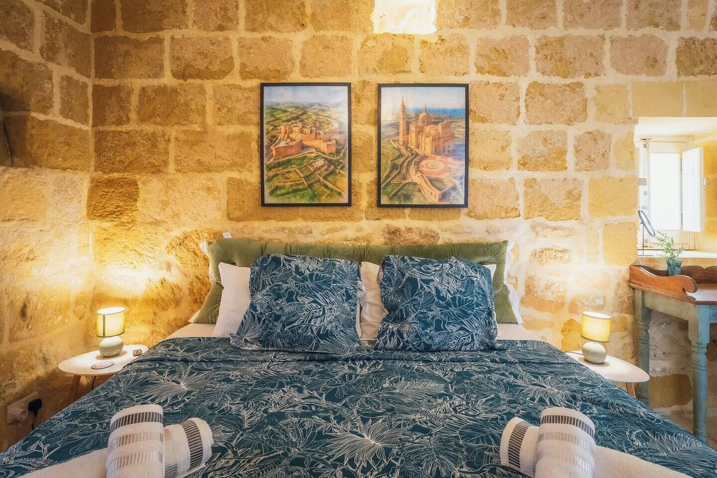 A bedroom with a stone wall, a bed with a patterned duvet, two framed pictures, and bedside lamps.