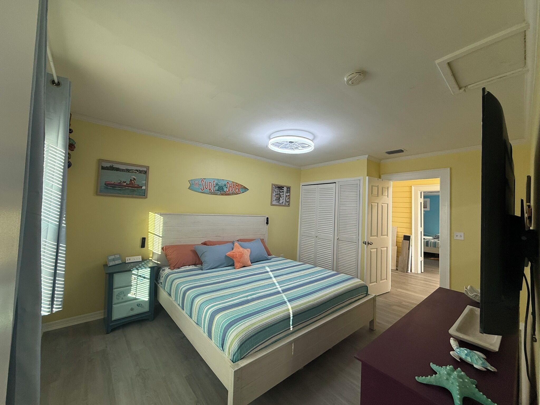 A bedroom with a bed, bedside table, and a wardrobe. There is a ceiling light and a door leading to another room.