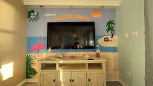 Huoneessa on seinälle asennettu televisio, sen alla puinen kaappi sekä seinämaalaus, jossa on kuvattuna trooppinen maisema: flamingo, palmu ja ranta.