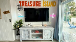 A living room with a large flat-screen TV mounted on the wall, a white cabinet with decorative items, a potted plant, and a sign reading "TREASURE ISLAND".