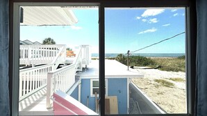 Vista desde una habitación con un ventanal grande que da a una playa, con barandilla blanca y un edificio azul al fondo.