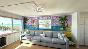 Una habitación con un sofá gris, un mural con escena de playa y una ventana con vista al océano.