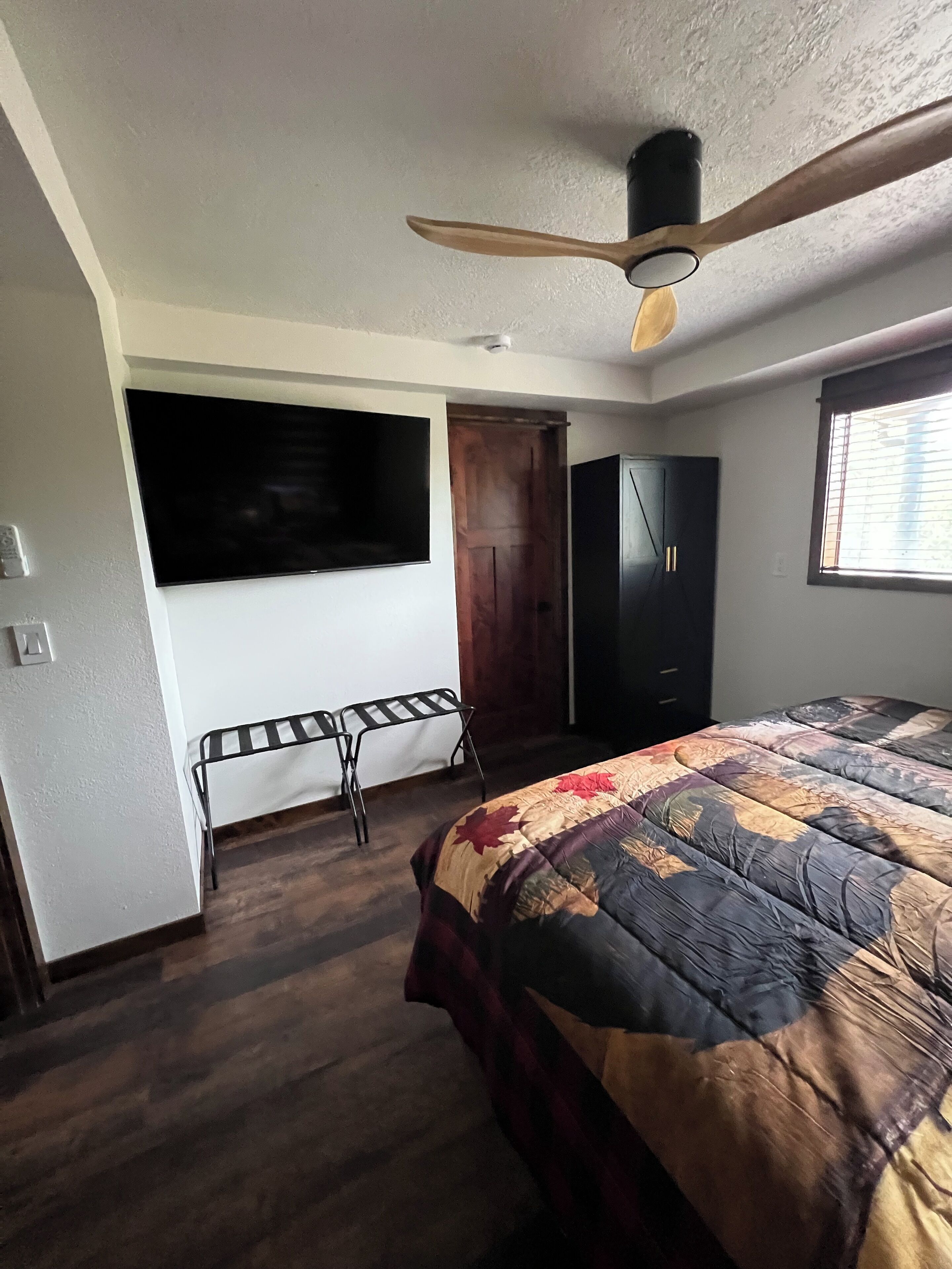 Een slaapkamer met een bed, een plafondventilator, een kledingkast, een aan de muur bevestigde televisie en een raam.