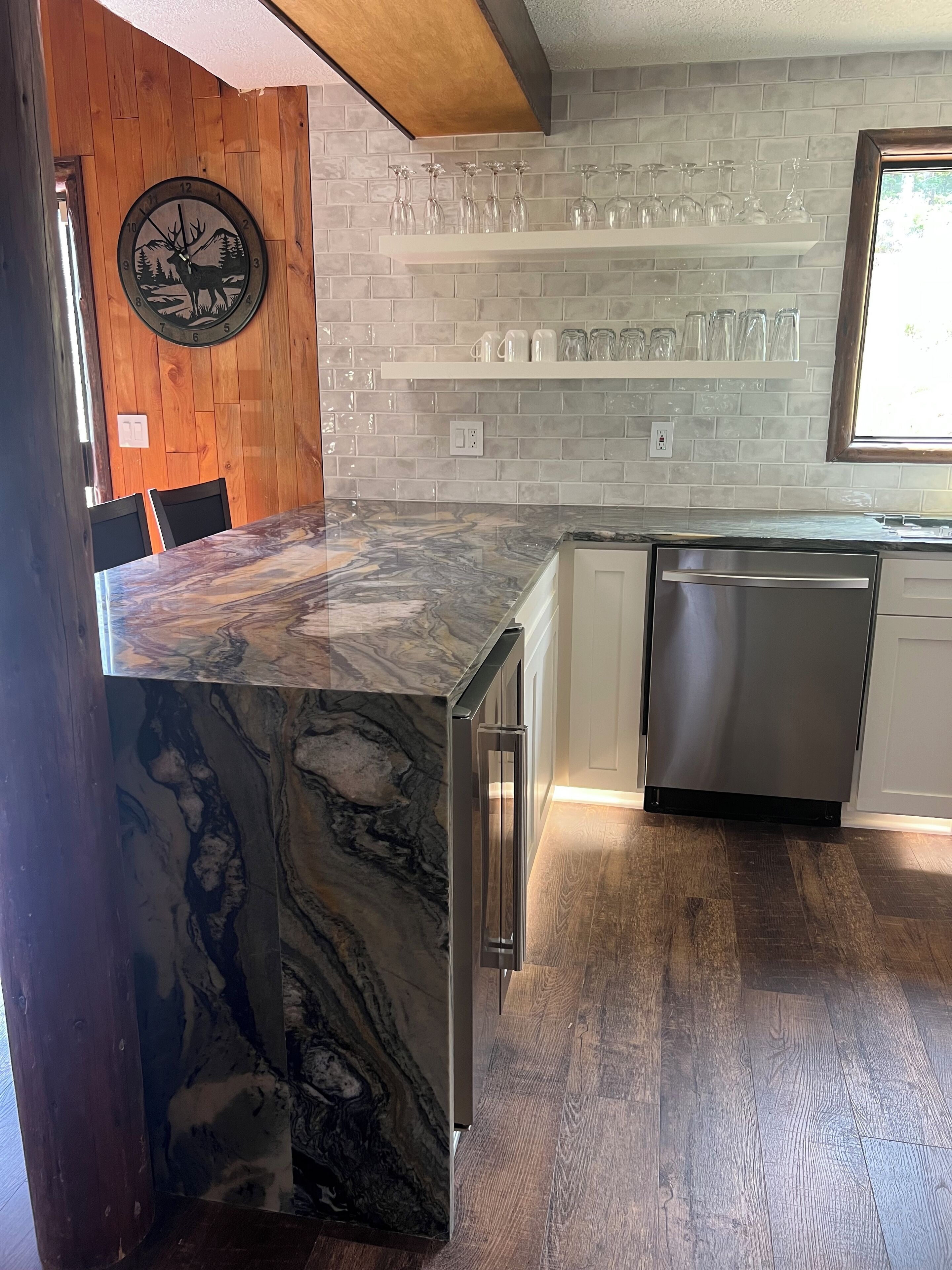A kitchen with a marble countertop, stainless steel dishwasher, and a brick backsplash.