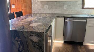 A kitchen with a marble countertop, stainless steel dishwasher, and a brick backsplash.