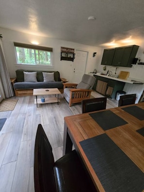 A compact living space with a sofa, a dining table, a kitchen area, and a window with a view of greenery. - Moss Top Cabin-off grid living moss roof cabin in the woods (Marblemount)
