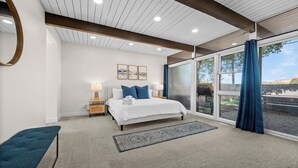A bedroom with a large bed, a nightstand, a bedside table, a blue bench, a large window with a view, and a ceiling with wooden beams and recessed lighting.