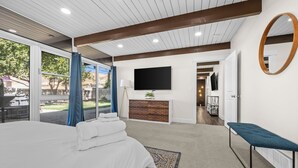 A modern hotel room with a large bed, a flat-screen TV mounted on the wall, a wooden ceiling, and a view of the outdoors.