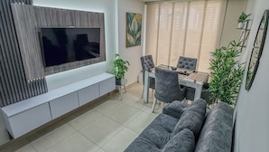 Een moderne woonkamer met een grijze bank, een flatscreen televisie bevestigd aan een houten paneel, een eettafel met stoelen, en een potplant.