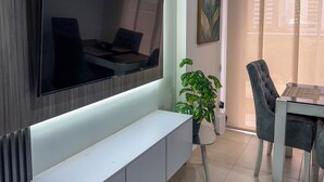 Een moderne woonkamer met een grote flatscreen-tv aan de muur, een witte console tafel, een eethoek met een tafel en stoelen, en een potplant.