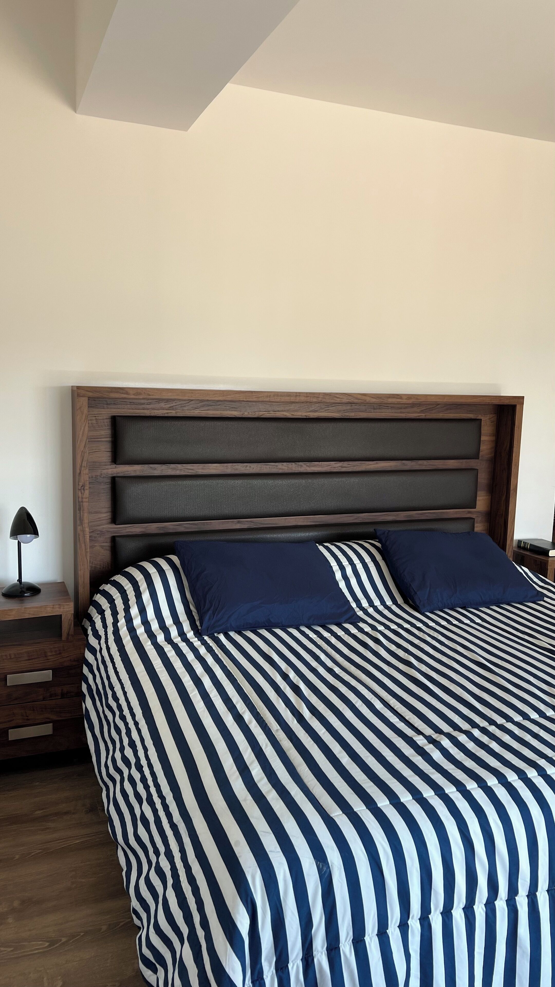 Una cama con cubrecama a rayas y dos almohadas, un cabecero de madera, una mesita de noche con una lámpara y un libro.