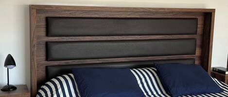 Una cama con cubrecama a rayas y dos almohadas, un cabecero de madera, una mesita de noche con una lámpara y un libro.