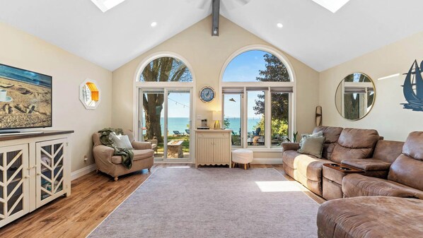 A spacious living room with a high ceiling, large windows offering a view of the sea, a cozy seating area with a fireplace, and a wooden cabinet with glass doors.