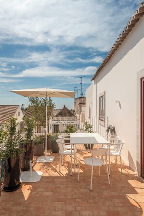 Un patio al aire libre con una mesa y sillas, rodeado de plantas en macetas, bajo un paraguas grande.