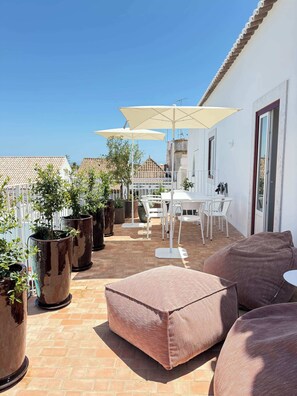 Un patio con una mesa y sillas blancas, plantas grandes en macetas y un paraguas color beige.