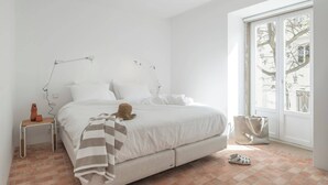 A minimalist bedroom with a large bed, a bedside table, a lamp, and a view of the outdoors through a window. - Danna Portuguese Design House nº 10 (Tavira)
