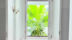 Quarto com janela que dá para uma bananeira grande, um banco de madeira com almofadas vermelhas e um chão de tijolos rústicos.