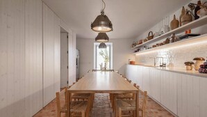 Een lange houten eettafel met stoelen, een hanglamp, witte kasten en een vloer van baksteen.